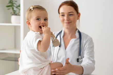 Baby playing with wooden toy while a woman wearing a white lab coat and stethoscope around her neck stands smiling in the background