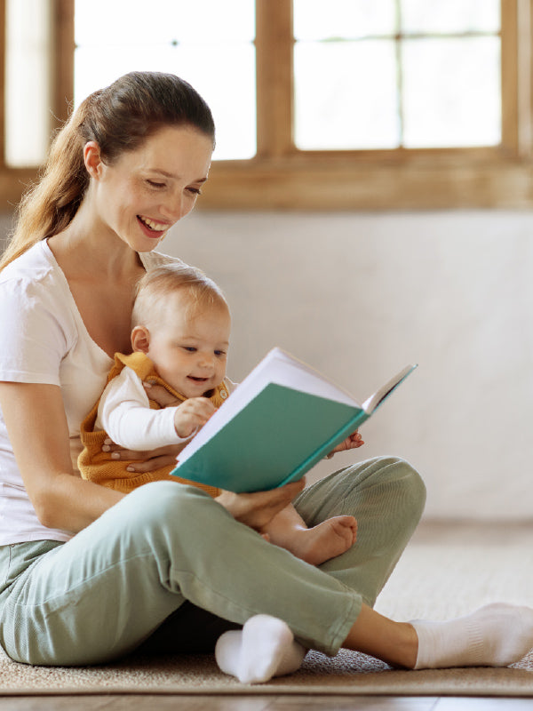 A woman sitting on a carpeted floor reads a book to a baby in a bright, softly lit room with a window in the background.